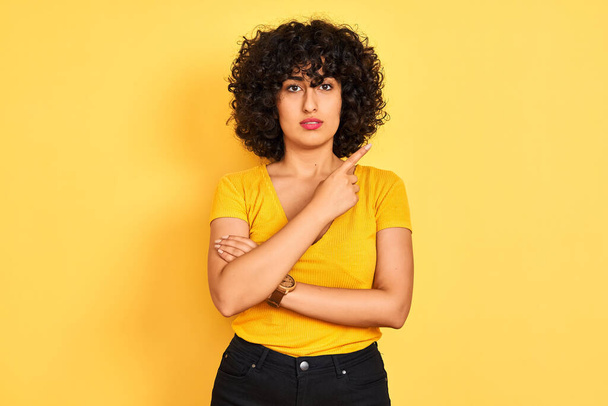 Молодая арабская женщина с кудрявыми волосами в футболке стоит на изолированном желтом фоне указывая пальцем руки в сторону показывая рекламу, серьезное и спокойное лицо
 - Фото, изображение