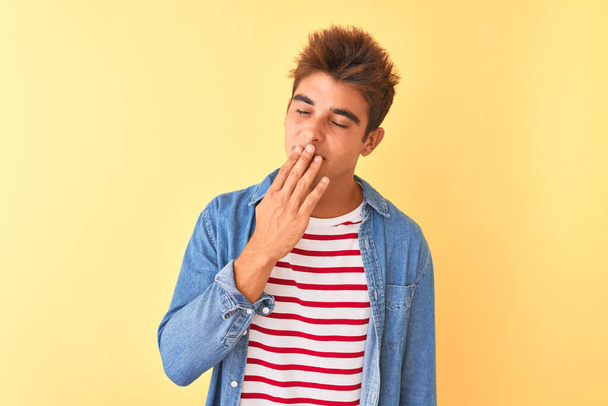 Jonge knappe man draagt gestreept t-shirt en denim shirt over geïsoleerde gele achtergrond verveeld gapen moe bedekken mond met de hand. Rusteloosheid en slaperigheid. - Foto, afbeelding