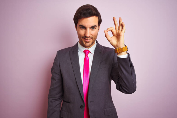 junger gutaussehender Geschäftsmann in Anzug und Krawatte, der vor isoliertem rosafarbenem Hintergrund steht und positiv lächelt und Ok-Zeichen mit Händen und Fingern macht. Erfolgreicher Ausdruck. - Foto, Bild