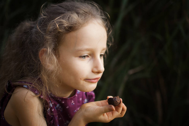 Mooi blond meisje in violette jurk met paardenstaart is het eten van een chocolade snoep in het park durring een dag, suiker verslaving concept - Foto, afbeelding