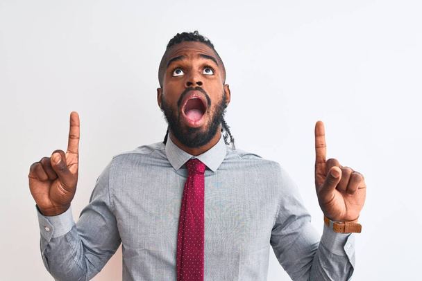 Afrikaans-Amerikaanse zakenman met vlechten dragen stropdas staande over geïsoleerde witte achtergrond verbaasd en verrast omhoog te kijken en te wijzen met vingers en opgeheven armen. - Foto, afbeelding