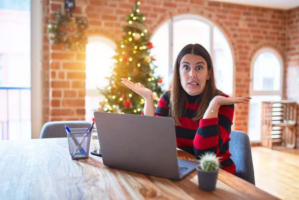 Krásná žena sedící u stolu a pracující doma s laptopem kolem vánočního stromku bezradný a zmatený výraz s rukama a rukama zdviženýma. Pochybný koncept. - Fotografie, Obrázek