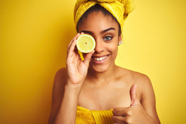 Αφρογυναίκα φορώντας πετσέτα μετά το ντους κρατώντας φέτα λεμόνι πάνω από απομονωμένο κίτρινο φόντο χαρούμενο με μεγάλο χαμόγελο κάνει OK πινακίδα, αντίχειρας με τα δάχτυλα, εξαιρετική πινακίδα - Φωτογραφία, εικόνα
