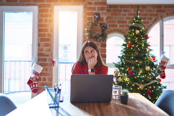 クリスマスツリーの周りの自宅でノートパソコンを使って作業テーブルに座っている美しい女性は、口の爪をかむ上で手でストレスと緊張しています。不安の問題. - 写真・画像