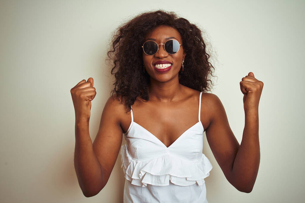 Young african american woman wearing t-shirt and sunglasses over isolated white background celebrating surprised and amazed for success with arms raised and open eyes. Winner concept. - Photo, Image