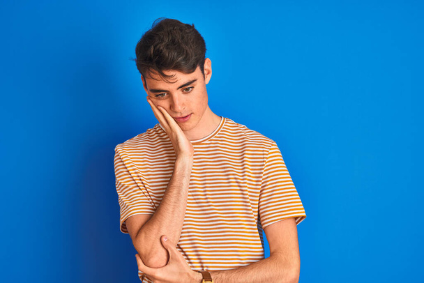 Teenager-Junge in lässigem T-Shirt, der über blauem, isoliertem Hintergrund steht und müde und gelangweilt mit verschränkten Armen aussieht. - Foto, Bild