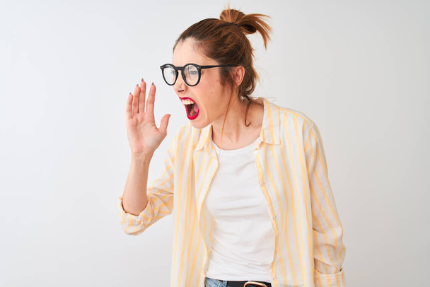 Zrzavá žena v pruhované košili a brýlích, stojící nad osamělým bílým pozadím, křičící a křičící na stranu s rukou na ústech. Koncept komunikace. - Fotografie, Obrázek