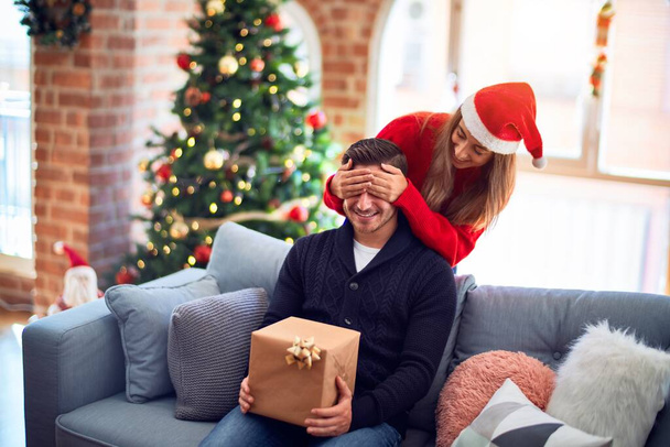 Молодая красивая пара улыбается счастливой и уверенной. Женщина, закрывающая глаза мужчине, окружает ее подарком вокруг рождественской елки дома
 - Фото, изображение