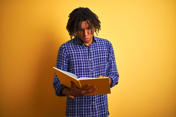 Афро-американский студент с дредами, читающий книгу на изолированном желтом фоне, напуганный от удивления лицом, напуганный и взволнованный выражением страха
 - Фото, изображение