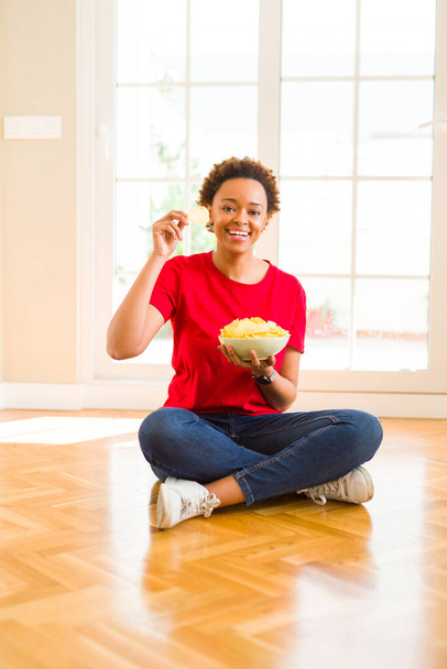 Mladá krásná africká americká žena s afro vlasy jíst chipsy sedí na podlaze doma - Fotografie, Obrázek