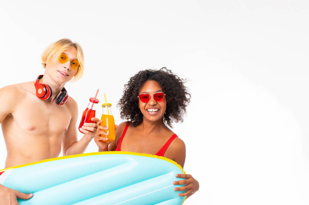 African woman and Caucasian blonde man in swimwear posing with air mattress and beverages against white background  - Фото, изображение