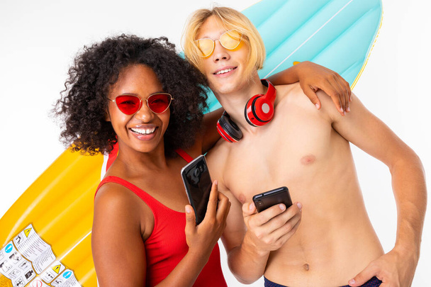 African woman and Caucasian blonde man in swimwear posing with mobile phones against air mattress - Photo, Image