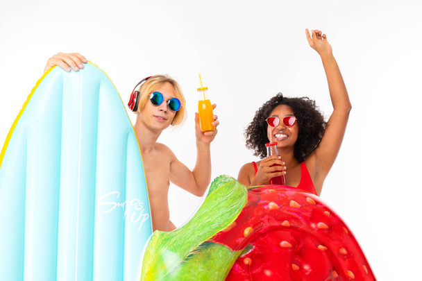 African woman and Caucasian blonde man in swimwear posing with air mattress and beverages against white background  - 写真・画像