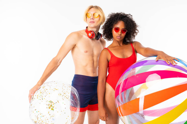 African woman and Caucasian blonde man in swimwear posing with rubber balls against white background  - Foto, Bild