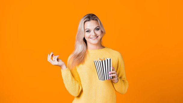 Young cheerful woman in a yellow sweater watching a movie and eating popcorn. - Photo, Image