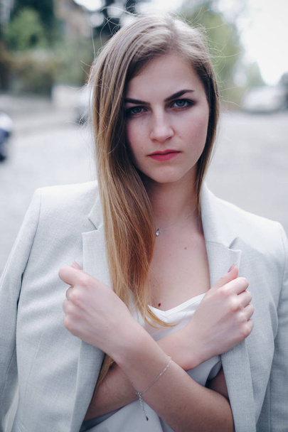 Portrait pf young caucasian woman with long blonde hair, soft light skin, make up, birthmarks and silver accessories looking at camera. Female student in white stylish suit crossing her arms and holding jacket. Concept of fashion, education and busin - Foto, imagen