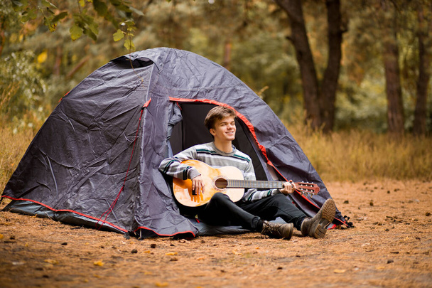 Glimlachende jongeman die in de buurt van een toeristische tent zit en gitaar speelt in het herfstbos - Foto, afbeelding