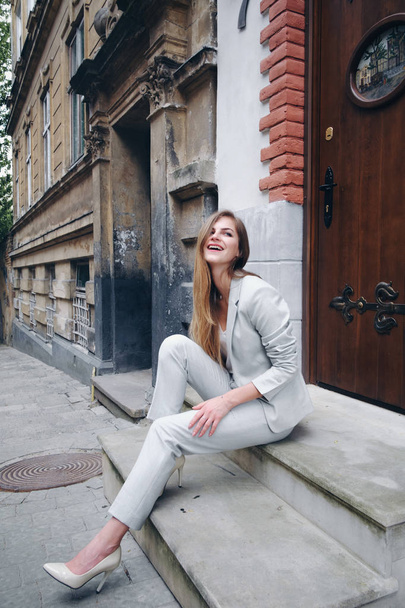 Side view of pretty girl, dressing in stylish white suit and high heels,sitting on steps near old wood door. Beautiful woman laughing and looking away. Concept of beauty. - Fotoğraf, Görsel