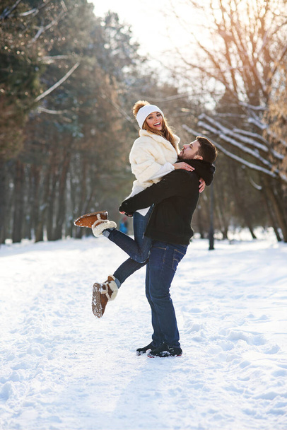 Young stylish couple in love have fun while walking on the snowy winter park. Handsome young man holds his beautiful girl on the hands. - Foto, Imagem