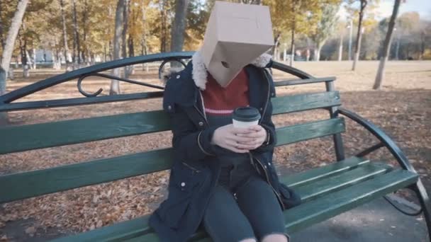 La fille avec le paquet sur la tête assise sur un banc avec une tasse de café. Fille dans une veste - Séquence, vidéo