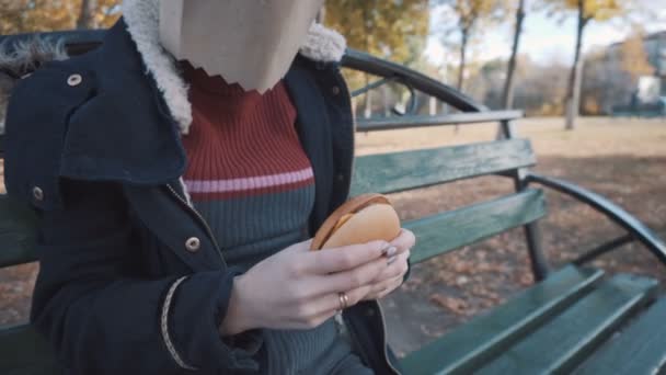 La fille avec le paquet sur la tête est assise sur un banc avec un cheeseburger. Fille dans une veste - Séquence, vidéo