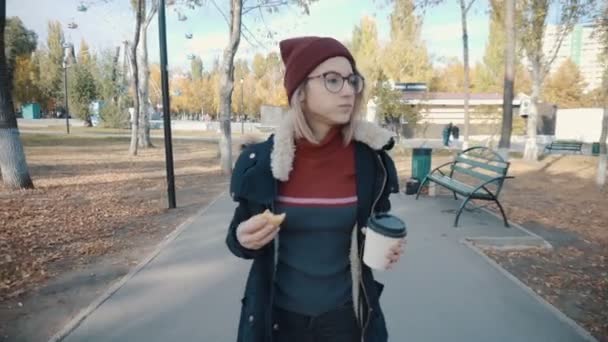 Girl walks in the Park and eating a burger and drink coffee.Girl in a jacket and hat - Imágenes, Vídeo