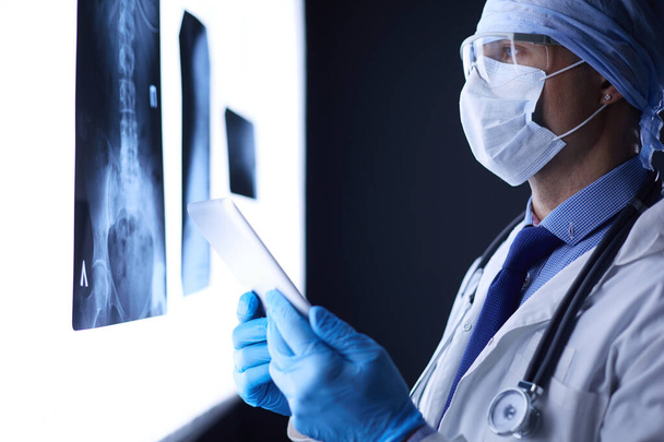 Doctor in hospital sitting at desk looking at x-rays on tablet against white background with x-rays - Foto, Imagem