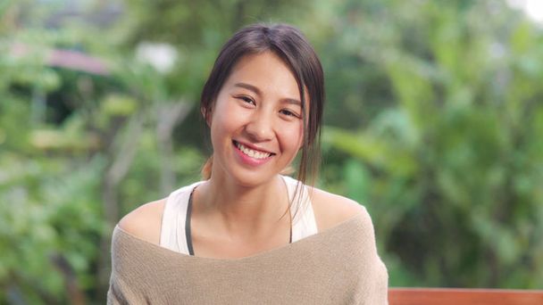 Aasialainen nainen hymyilee onnellisena ja etsii kameraa rentoutuessaan pöydällä puutarhassa kotona aamulla. Elämäntapa naiset rentoutua kotona käsite.  - Valokuva, kuva