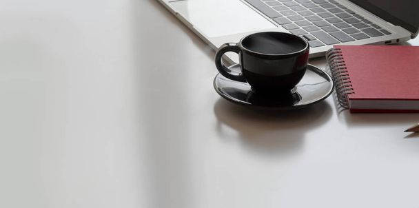 ノートパソコンと白の机の背景にオフィス用品と黒のコーヒーカップと現代のワークスペースの作物のショット - 写真・画像