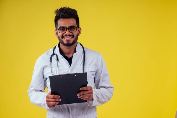 indian male doctor wearing glasses and white cloakholding tablet in hand in yellow background studio - Фото, зображення