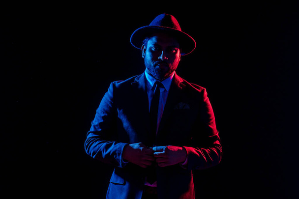 Young handsome model posing in a studio in a trendy neon light in a stylish suit and hat - Foto, Imagem
