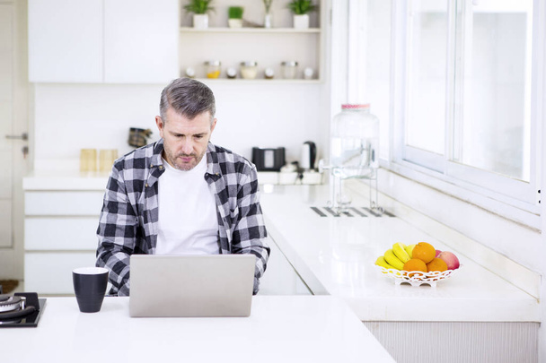 Mature man working with a laptop in the kitchen - Photo, image