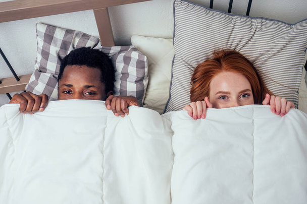 rossiccio zenzero caucasico felice femminile e multi-etnico afro uomo insieme sdraiato nella camera da letto letto nascondersi sotto il concetto blanket.bashfulness
 - Foto, immagini