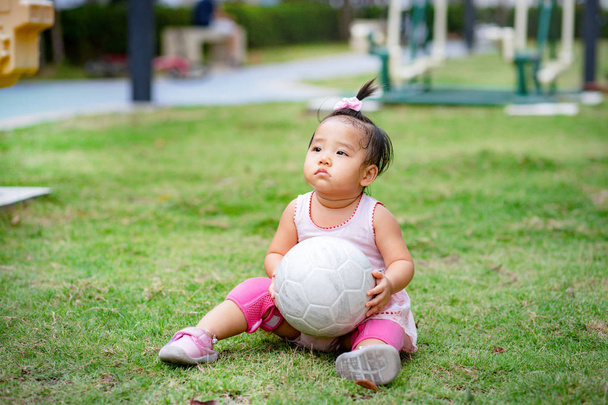 Niña sentada en el campo de hierba y sosteniendo la pelota en el parque
 - Foto, Imagen