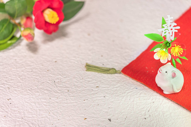 Μια ιαπωνική ευχετήρια κάρτα με ένα λουλούδι Tsubaki που ονομάζεται επίσης το χειμερινό τριαντάφυλλο και ένα χαριτωμένο ειδώλιο αρουραίου για το 2020 έτος του ποντικιού σε ένα κόκκινο πανί διακοσμημένο με την ιαπωνική ιδεογραφία Kotobuki που σημαίνει μακροζωία σε ένα washi τσαλακωμένο χαρτί - Φωτογραφία, εικόνα
