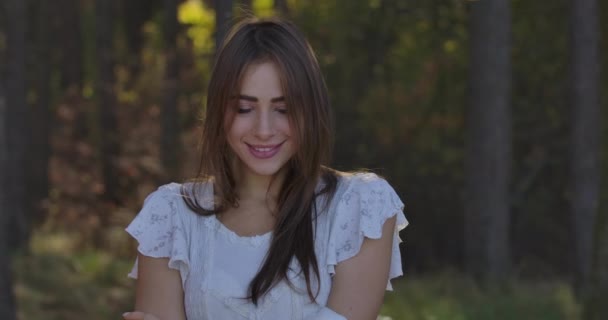 Portrait of a Caucasian brunette woman with brown eyes standing between trees in the summer forest. Charming girl in white dress posing to the camera and smiling. - Video