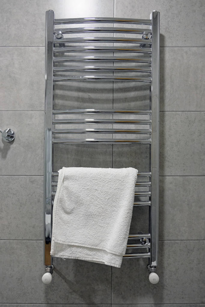 Roestvrij elektrische handdoekdroger voor de badkamer. Drogen is gemonteerd op een muur met grijze tegels, witte handdoek. Handdoekdroger voor het drogen van handdoeken in de badkamer, verwarming en drogen van de badkamer - Foto, afbeelding