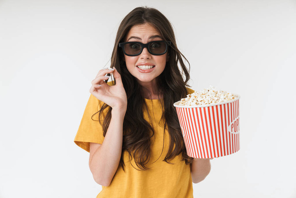 verwarde jonge vrouw geïsoleerd over witte muur achtergrond in 3D bril eten popcorn horloge film. - Foto, afbeelding