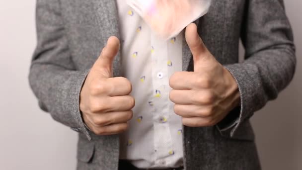 Solid caucasian man in gray jacket and shirt on white background pulls approving thumbs up sign. Money banknotes falling around him. wealth, richness and wasting dough. Bills tossing, spending money - Footage, Video
