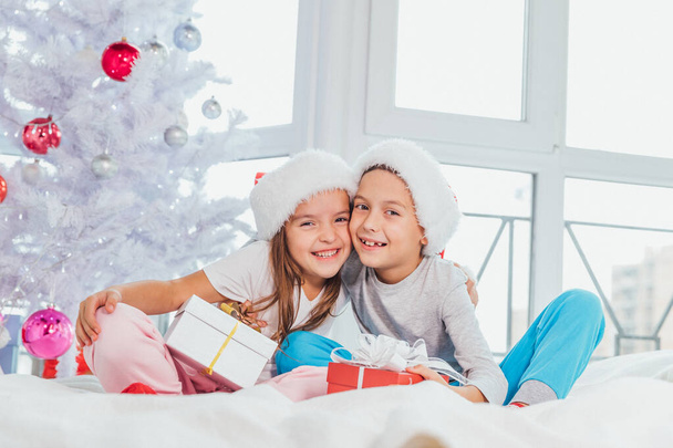 Image of brother and sister in santa hats, tenderly embracing, sharing Christmas presents, sitting on the white bed linen, which looks like a snow. - Fotografie, Obrázek