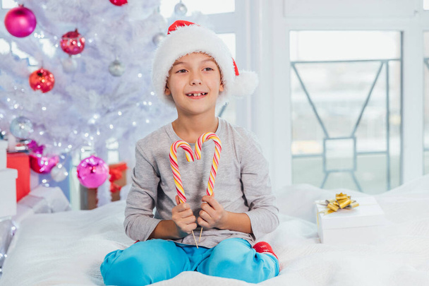 Funny boy in santa hat is sitting on the bed in pyjamas, demonstrating striped candy canes. - Fotoğraf, Görsel