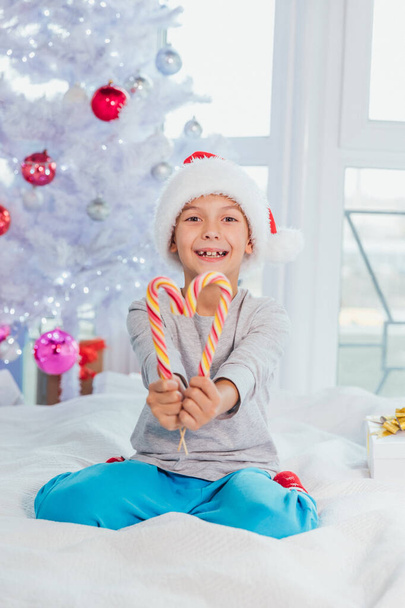 Funny boy in santa hat is sitting on the bed in pyjamas, demonstrating striped candy canes. - Φωτογραφία, εικόνα