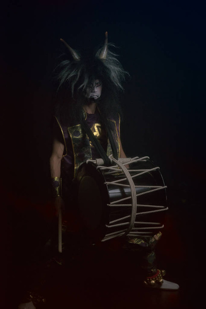 Demon from japanese mythology full lenght portrait in a darkness. Taiko drummer in a wig and a demon mask sitting and smiling on black background. - Photo, Image
