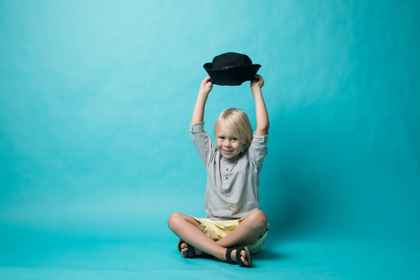 Un niño sobre un fondo azul se sienta en un sombrero negro y mira a la cámara. El chico lleva una camiseta gris. Feliz.
 - Foto, imagen
