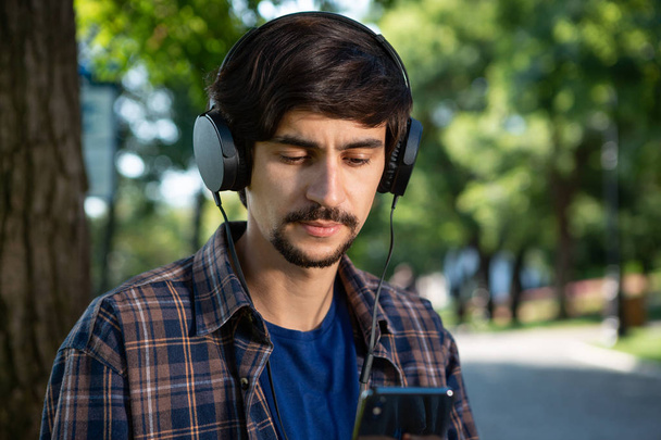 Handsome brunet bearded man in a check shirt listening to music on a smartphone enjoying warm day in a park. - Φωτογραφία, εικόνα