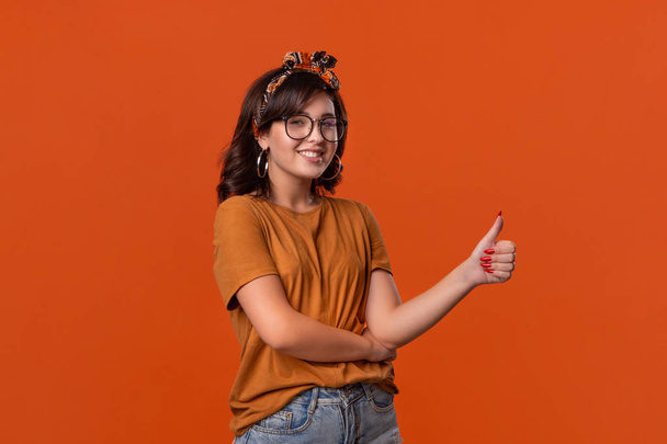 Uśmiechnięta brunetka w pomarańczowym t-shircie, opasce i okularach trzymająca kciuki w górze wyrażająca pozytywną ocenę stojącą na pomarańczowym tle. - Zdjęcie, obraz
