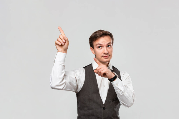 Przystojny mężczyzna w białej koszuli i szarej kamizelce pokazuje palcami pustą przestrzeń do reklamy w lewym górnym rogu jasnoszarego tła. - Zdjęcie, obraz
