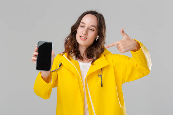 グレーの背景に隔離された携帯電話の画面を示す黄色のレインコートで興奮したブルネットの女性。モデルはワイヤレスイヤーバッド. - 写真・画像