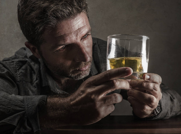alcoholic depressed and wasted addict man sitting in front of whiskey glass trying holding on drinking in dramatic expression suffering alcoholism and alcohol addiction - Foto, afbeelding