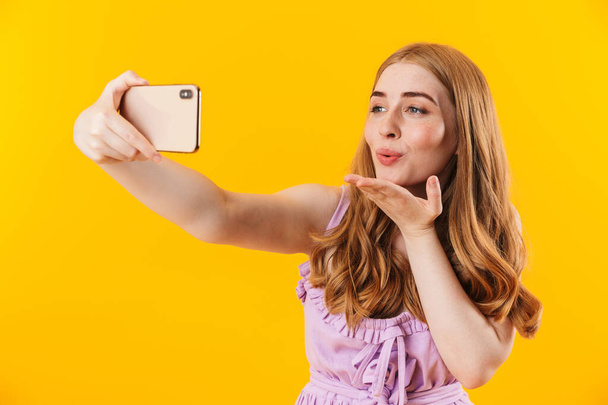 Cute pleased girl isolated over yellow wall background take selfie by mobile phone blowing kisses. - Fotografie, Obrázek